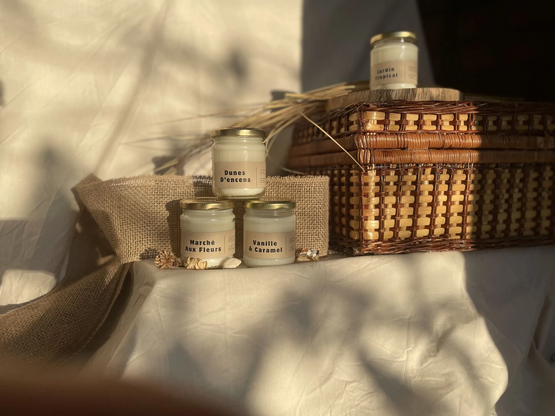 Bougies parfumées artisanales présentées sur un panier en osier. ougies artisanales parfumées disposées sur un panier en osier, avec des parfums comme Dunes d'Encens et Vanille & Caramel.